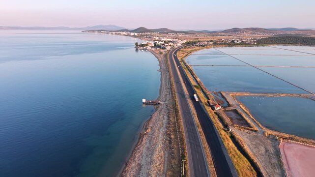 Aerial view of coastal salt evaporation ponds beside a highway and calm sea, showing pink and blue saline pools, shoreline infrastructure, farmland and distant mountains at sunset.
