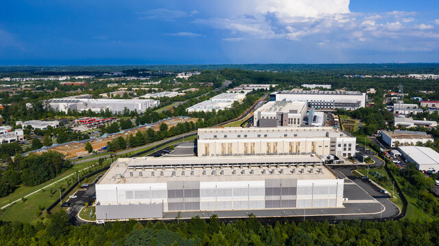 Aerial view of a large data center complex with white rooftops, cooling units, and surrounding green landscape under a blue sky in Ashburn, Virginia, United States.