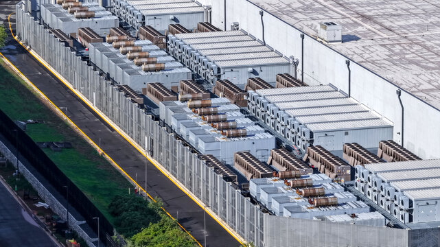 Aerial view of a large data center facility in Ashburn, featuring rows of industrial cooling units and HVAC systems on the roof, surrounded by a fence. Ashburn, Virginia, United States.