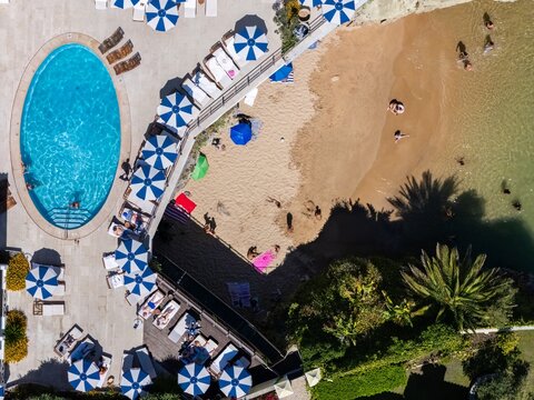 Aerial view of a beach resort featuring a blue swimming pool, sandy beach, and rows of blue and white striped umbrellas with people relaxing in Cascais, Portugal.