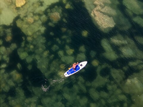 Aerial view of a person on a standup paddleboard gliding over clear shallow water with visible rocky seabed in Cascais, Portugal.