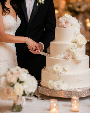 Bride and groom cut a tiered ivory wedding cake decorated with blush flowers, captured in warm reception light with soft candle and floral bokeh.