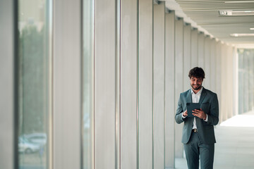 Fototapeta premium Professional businessman walking in modern office hallway, smiling and using digital tablet for work communication