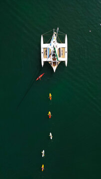 Aerial view of a catamaran anchored near Pointe du Cosmeur with a line of colorful kayaks paddling in the deep green water of Carantec, Bretagne, France.