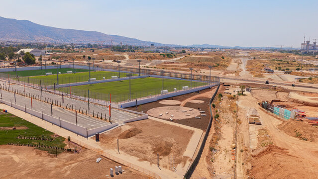 Aerial view of soccer fields under construction in a large development area with dirt ground, fencing, and stadium lights in Athens, Greece.