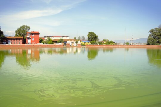 Scenic view of Siddha Pokhari, a historic pond with green water, reflections, traditional buildings and distant mountains in Bhaktapur, Nepal