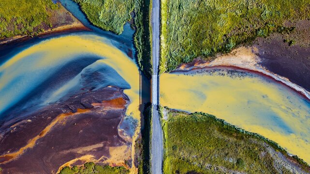 Aerial view of Hrutsvatn bridge crossing a river with vibrant yellow and blue water surrounded by lush green landscape in Borgarbyggd, Iceland.