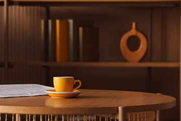 Yellow coffee cup and saucer on a wooden table with a newspaper and blurred bookshelf in the background, the background features a blurred bookshelf with books and decorative items © Pixel-Shot