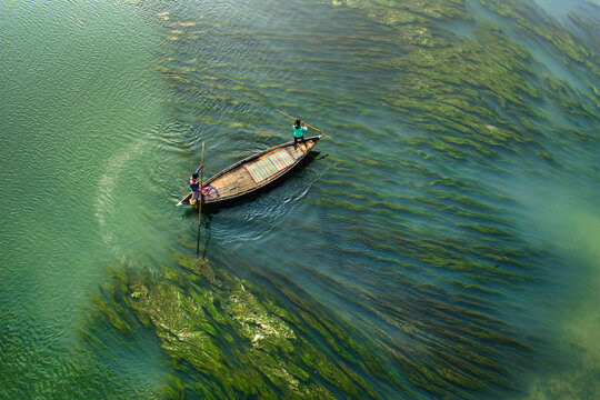 Aerial view of two fishermen in a traditional wooden boat navigating through lush green aquatic plants in the river in Sirajganj, Rajshahi Division, Bangladesh.