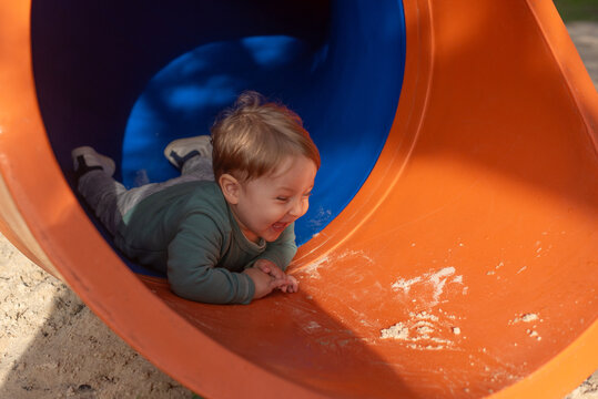 A happy 20-month-old toddler boy smiling as he goes down a covered playground slide at a sandy playground