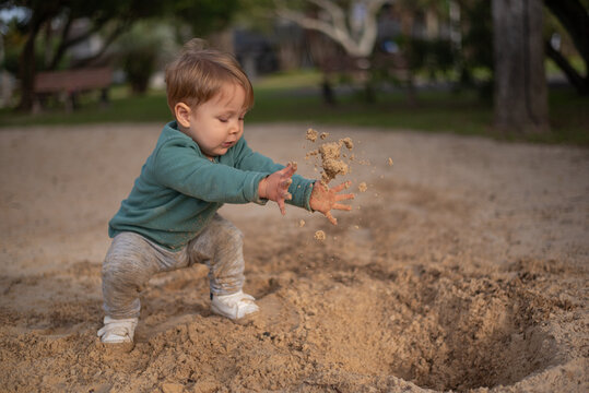 A 20-month-old toddler boy playing in the sand, throwing a handful of sand into a hole