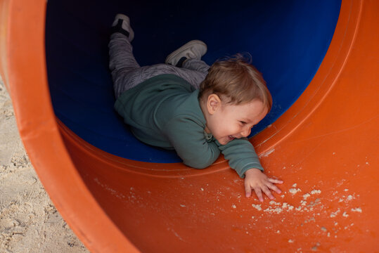 A happy 20-month-old toddler boy smiling as he goes down a covered playground slide at a sandy playground