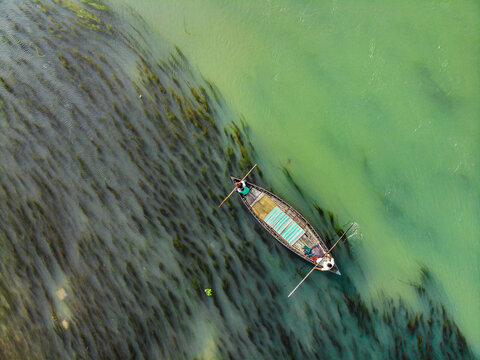 Aerial view of a small wooden boat with two people rowing on a river with visible underwater vegetation in Sirajganj, Rajshahi Division, Bangladesh.