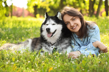 Happy young woman and her siberian husky dog lying together on green grass in a sunny park, the woman smiles at the camera while the dog rests beside her with its tongue out © Pixel-Shot