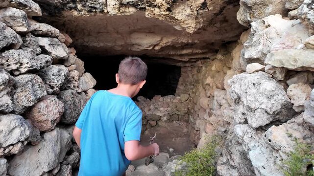 Young boys explores ancient stone cave. He bends to examine ancient stone structure. Darkness surrounds him, lit only by his flashlight. History whispers in ancient, echoing stone walls