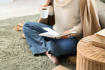 Obraz premium Young woman with book and cup of hot coffee sitting on carpet in living room