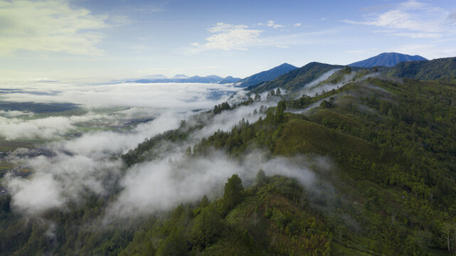 Aerial view of the lush, mist-covered mountains featuring dense green forests, rolling hills, and low-hanging clouds under a bright sky in Kerinci Regency, Jambi, Indonesia.