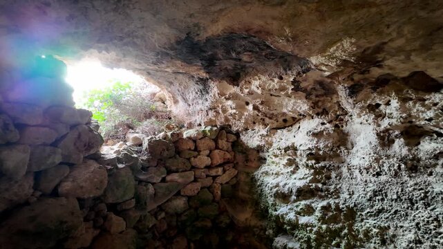 Sunlight pierces ancient stone cave entrance. Rough rock walls glow with natural light. Moss and lichen cover stone surfaces. Caves reveal history and natural beauty. Ideal for travel, heritage