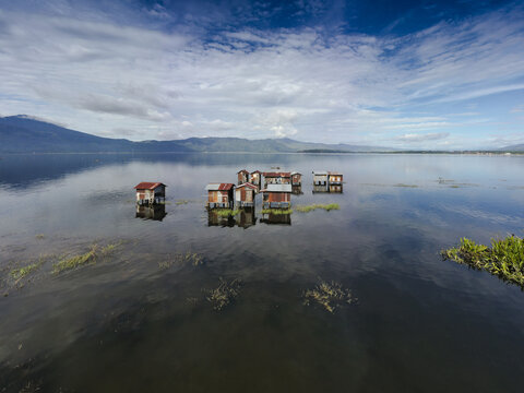 Aerial view of stilt houses on the calm waters of Lake Kerinci with distant mountains under a blue sky in Kerinci Regency, Jambi, Indonesia.