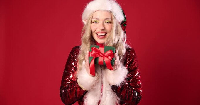 Woman dressed as elf looking around and showing Christmas gift on red background