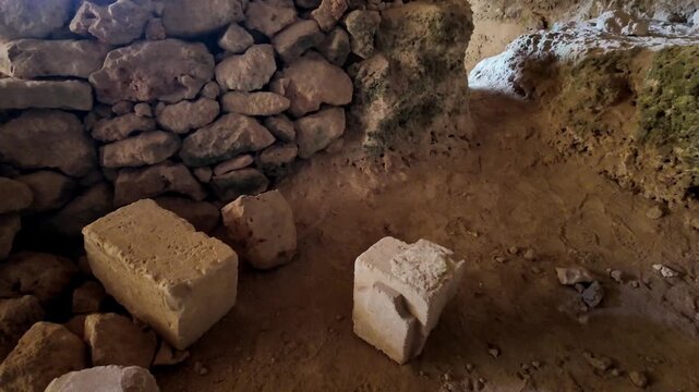 Ancient stone wall stands inside dimly lit cave. Large blocks lie on dirt floor nearby. Light filters through cave entrance above. Archaeological site reveals hidden history