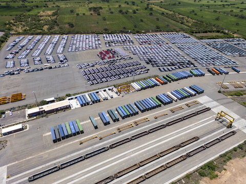 Aerial view of a car factory logistics yard with rows of new vehicles, colorful shipping containers, and freight train cars on tracks in San Luis Potosi, San Luis Potosi, Mexico.