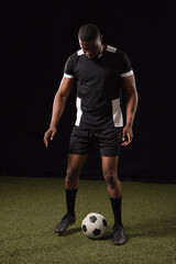 Fototapeta premium African American male standing on turf in studio wearing black-white soccer kit ball at feet