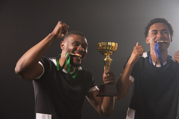 Fototapeta premium African male teammates celebrating in black-white jerseys, holding gold cup, biting medals in arena