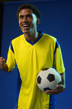 Adult African male wearing yellow jersey holding soccer ball clenching fist in studio
