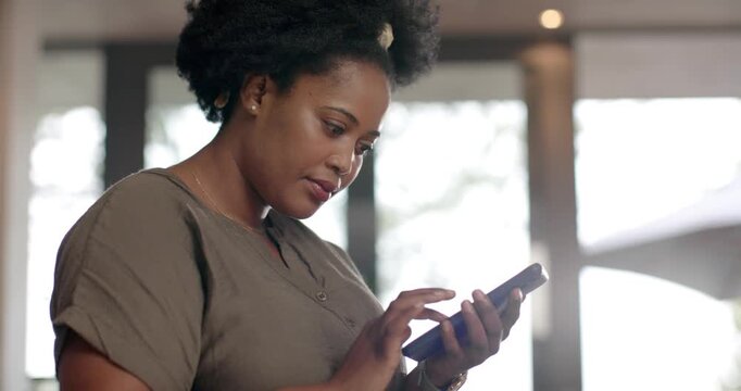 Adult African American woman reacting to message, tapping phone by tall windows, smiling, laughing
