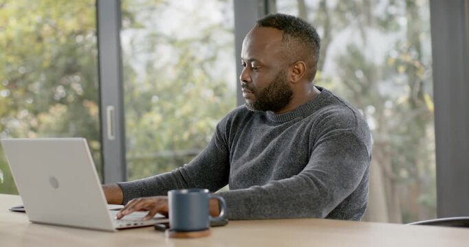 African man typing on laptop at table by glass doors pausing checking smartwatch clearing throat