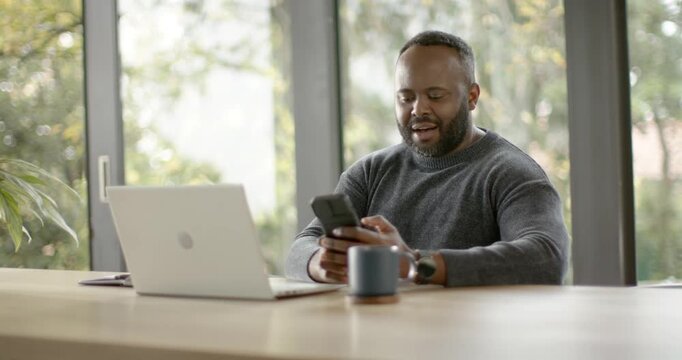 Adult African man receiving positive message, scrolling and cheering while typing on laptop at home