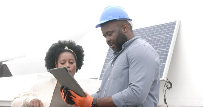 African American tech holding tablet and client reviewing on rooftop checking solar panel wiring