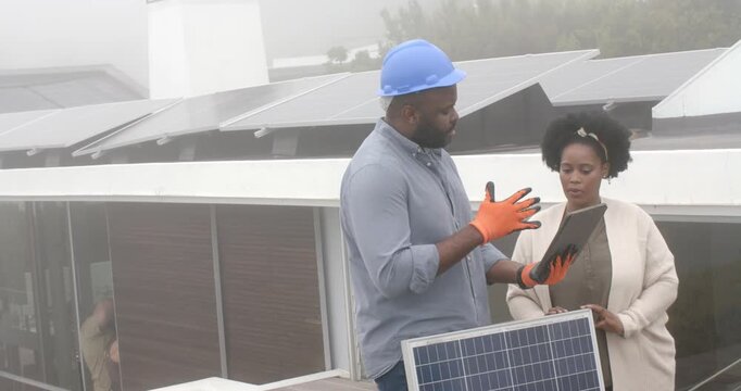 Technician in hardhat and client talking tablet showing panel data examining solar array on rooftop