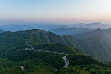 Fototapeta premium Layered green mountain ridges with wind turbines and a winding road, at dawn under a pale sky.