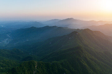 Fototapeta premium Layered forested mountain ranges stretch under a hazy sunrise sky, showing depth and natural landscape.