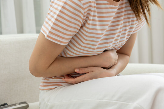 Woman stomach pain abdomen ache cramp sitting sofa home discomfort, woman holding abdomen while sitting sofa home, cropped view showing stomach pain, cramp, digestive discomfort, casual striped