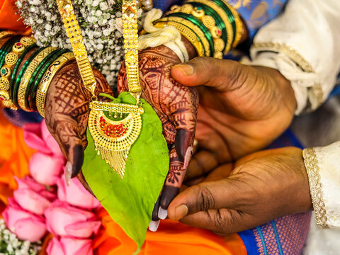 Indian Bride and Groom Hands Holding Betel Leaf During Wedding Ritual, rest on a fresh green betel leaf while holding a traditional gold necklace.