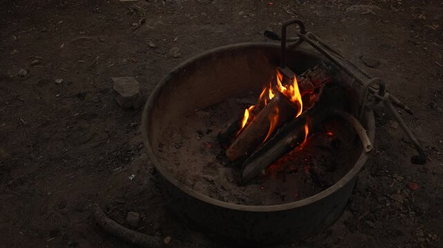 Burning logs glow inside metal fire pit at campsite, with orange flames, glowing embers and charred wood creating a warm evening campfire scene on the forest ground