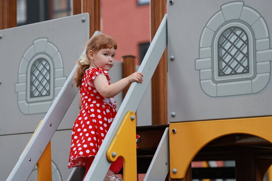A girl wearing a red dress with white spots is at the playground. She is looking closely at the slide while standing next to it. The area is lively with other children nearby.