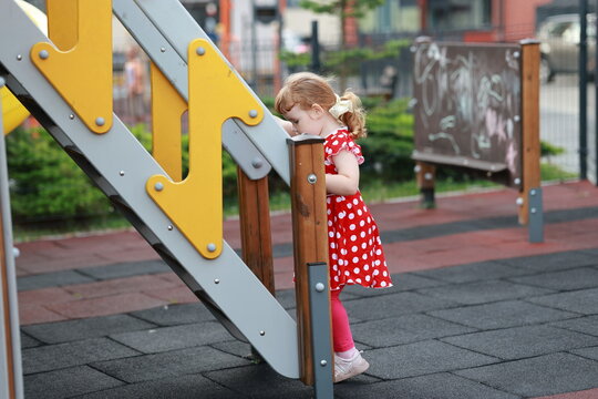 A girl wearing a red dress with white spots is at the playground. She is looking closely at the slide while standing next to it. The area is lively with other children nearby.