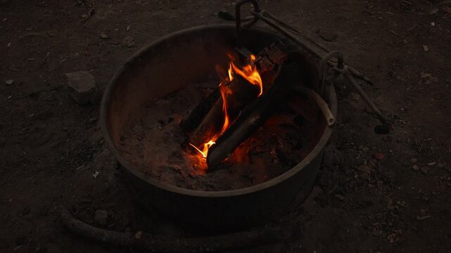 Burning logs glow with orange flames inside metal campsite fire pit, surrounded by ash and charred wood as the fire slowly burns in a dark outdoor camping area
