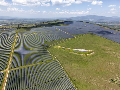 Aerial view of a large solar farm with rows of photovoltaic panels stretching across green fields and rolling hills under a blue sky in Nopala, Hidalgo, Mexico.