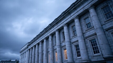 Fototapeta premium Low angle view of neoclassical building facade with tall columns and lit windows under dramatic cloudy evening sky