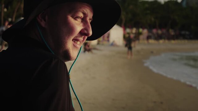 Closeup view of a young adult Caucasian man in black sun hat walking along the sea shore and talking to somebody. Slow motion of a smiling guy going at the beach and having conversation with a friend
