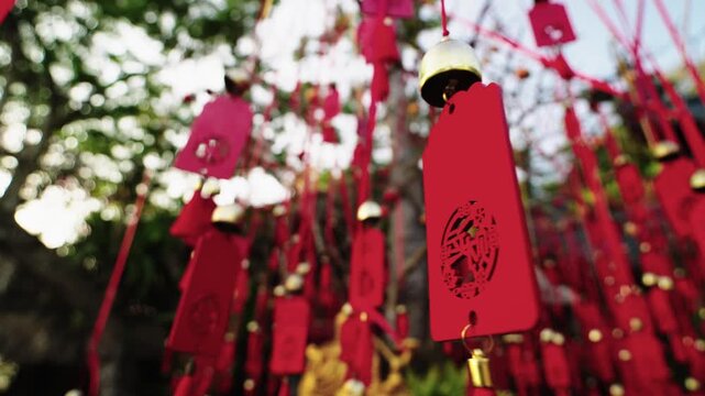 Lots of red wooden wish cards with bells are hanging on ribbons on tree branches moving by the wind filmed from above. Many Buddhist tablets with holy desires are on plants in the park near temple