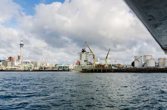 A detailed waterfront scene featuring a cargo vessel, industrial storage tanks, and Auckland&rsquo;s skyline rising in the background beneath soft, partly cloudy light
