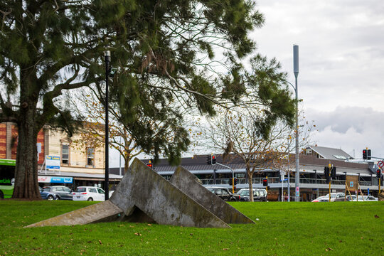 A bold angular concrete sculpture stands on the lawn at Western Park, framed by trees and the surrounding urban streets of Auckland
