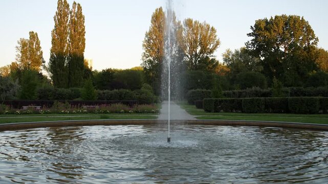 Slow pull back camera movement revealing a circular park fountain with a single jet shooting water upward, setting into a wide shot framed by lush trees illuminated at their tops by golden sunset ligh