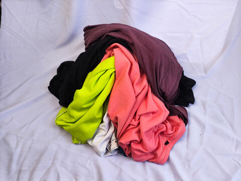 A messy pile of colorful t-shirts and cotton clothing, including bright pink, neon yellow, black, and maroon fabrics scattered on a clean white fabric surface.